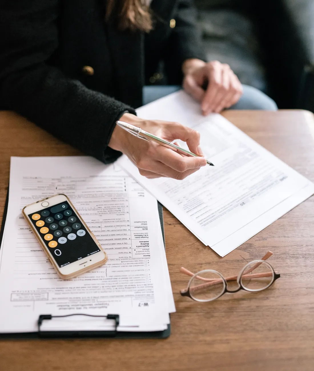 Tax forms, a phone calculator and reading glasses on a walnut desk as an accountant reviews a return